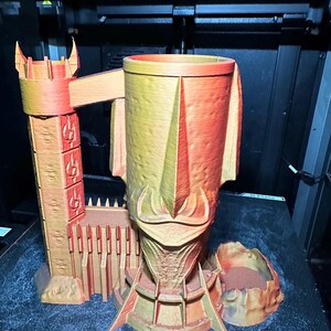 May include: A 3D printed, orange and yellow, castle-shaped mug with a tower and a wall. The mug is sitting on a black surface.