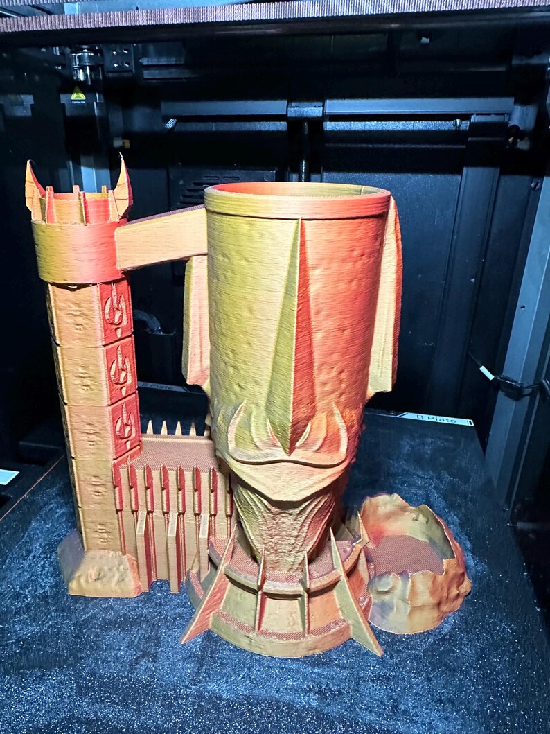 May include: A 3D printed, orange and yellow, castle-shaped mug with a tower and a wall. The mug is sitting on a black surface.