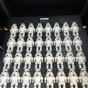 May include: A collection of white Stormtrooper figurines, arranged in rows on a black surface. The figurines are detailed with black accents and stand upright. The word "CREALITY" is visible on the surface.