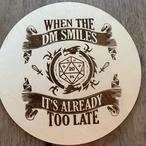 May include: A round wooden coaster with a brown and white design. The coaster features a 20-sided die, a dragon, and the text "When the DM smiles it's already too late".