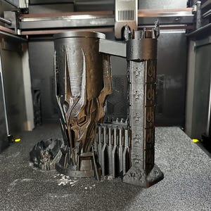 May include: A 3D printed black mug shaped like the tower of Barad-d&ucirc;r from the Lord of the Rings. The mug has a detailed design with the inscription "One Ring to rule them all" on the side.