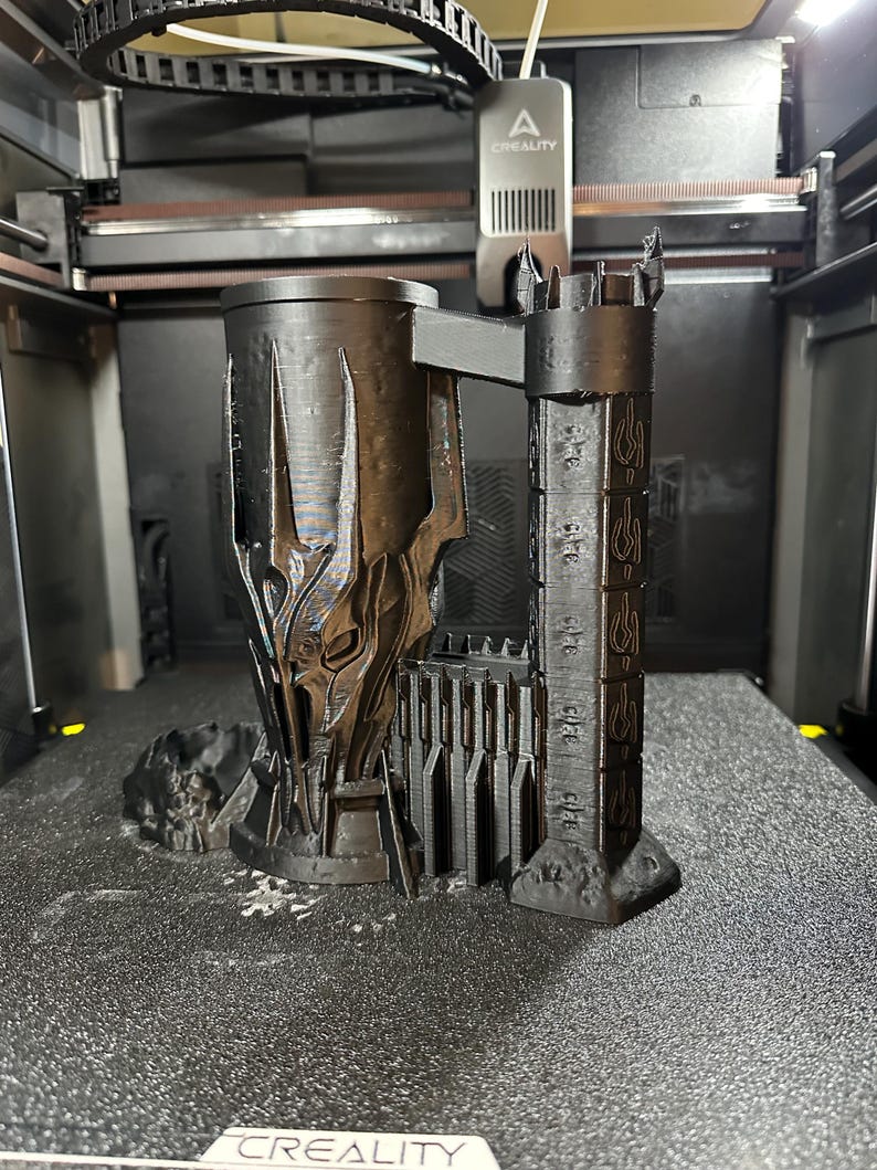 May include: A 3D printed black mug shaped like the tower of Barad-d&ucirc;r from the Lord of the Rings. The mug has a detailed design with the inscription "One Ring to rule them all" on the side.