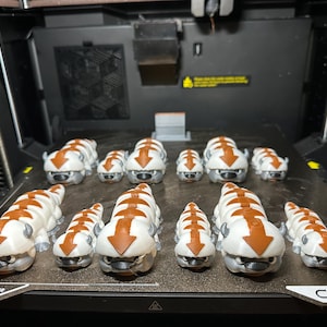 Op de afbeelding: Een verzameling wit en bruin 3D-geprinte figuren van Appa uit Avatar: The Last Airbender, gerangschikt op een 3D-printerbed. De figuren variëren in grootte, met grotere op de voorgrond en kleinere op de achtergrond.