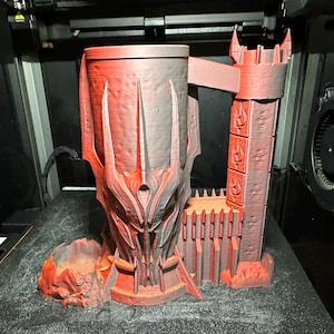 May include: A 3D printed red and grey mug shaped like a tower with a detailed design. The mug has a handle and a base with a small, separate piece that looks like a rock.