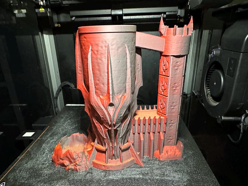 May include: A 3D printed red and grey mug shaped like a tower with a detailed design. The mug has a handle and a base with a small, separate piece that looks like a rock.