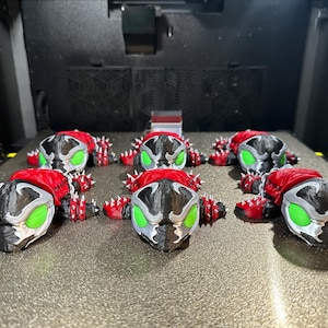 May include: Several 3D-printed figures of a comic book character with a red cape, black and silver mask, and green eyes. The figures are arranged on a textured surface inside a machine.