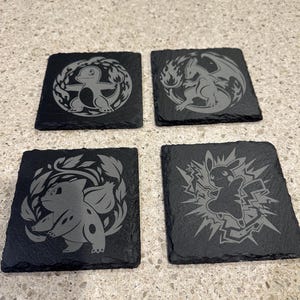May include: Four square black slate coasters with engraved Pokémon designs. Each coaster features a different character: Charmander, Charizard, Bulbasaur, and Pikachu. The designs are in a light gray color, set against the dark slate.