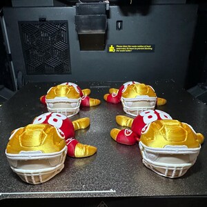 以下が含まれることがあります： 3Dプリンターで印刷された、金、赤、白のミニチュアのアメリカンフットボールヘルメット6個。ヘルメットには86の数字が入っています。ヘルメットは2列3列に配置されています。