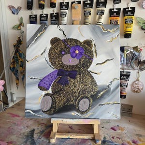 Könnte beinhalten: Quadratische Leinwandmalerei eines Teddybären mit lila Augenklappe und Blume. Der Bär hat braunes Fell, eine lila Fliege und einen lila Ärmel. Der Hintergrund ist grau mit goldenen und weißen Akzenten. Das Gemälde wird auf einer kleinen Holzstaffelei ausgestellt.