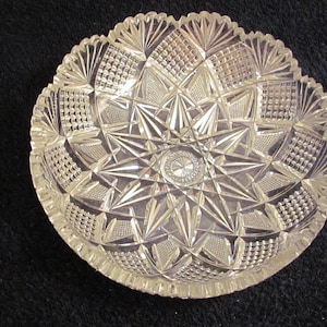 May include: Clear, cut-glass bowl with a starburst pattern in the center and a scalloped edge. The bowl's intricate design features geometric shapes and textures, reflecting light. A decorative piece for serving or display.