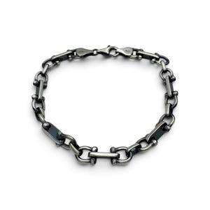 Könnte beinhalten: Ein dunkel silberfarbenes Metallkettenarmband mit einer Reihe von sich wiederholenden U-förmigen Gliedern, die durch flache rechteckige Stäbe verbunden sind. Das Armband ist kreisförmig angeordnet und hat einen sicheren Karabinerverschluss.