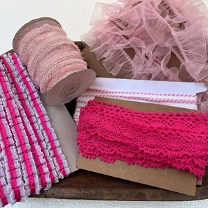 May include: A collection of pink and white ribbons and lace in a wooden bowl. Includes a spool of pink ribbon, pink lace trim, and pink and white striped ribbon. Perfect for crafting and sewing projects.