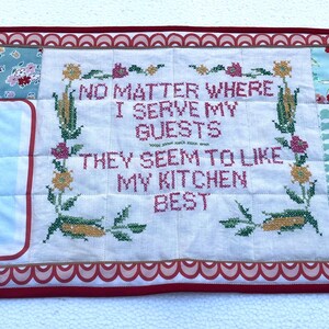 May include: A quilted table runner with a patchwork design. It features embroidered text that reads "NO MATTER WHERE I SERVE MY GUESTS THEY SEEM TO LIKE MY KITCHEN BEST." The runner has floral and geometric patterns and illustrations of women.