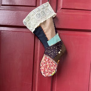 May include: A handmade Christmas stocking with a patchwork design. The stocking features various fabrics in shades of brown, blue, and red, with a white lace cuff. It is hanging against a red door.