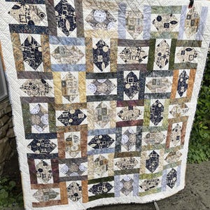 May include: A patchwork quilt with a repeating pattern of clock faces and geometric shapes in shades of beige, blue, and brown. The quilt is bordered with a white fabric and has a quilted texture.