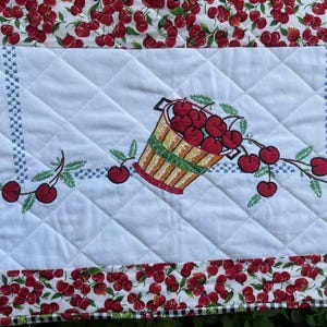 May include: A quilted fabric piece featuring a central white panel with a diamond-stitched pattern. The center displays an embroidered basket filled with red cherries. The border consists of red and white striped fabric and cherry-patterned fabric.