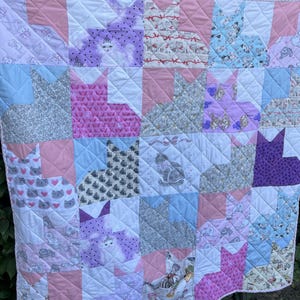 May include: A quilted blanket featuring a patchwork design of cat-shaped blocks. The fabric patterns include various illustrations of cats, kittens, bows, and hearts in shades of pink, blue, purple, gray, and white, all stitched with a diamond pattern.