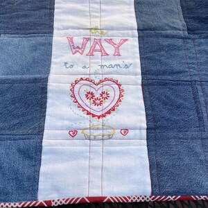 May include: A denim quilt with a white center panel featuring embroidered text and a heart design. The text reads "the WAY to a man's". The heart is red with floral details. The quilt has a red and white patterned border.