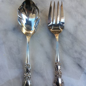 May include: A silver serving set with a spoon and fork. The spoon has a decorative, scalloped edge and the fork has a traditional design. Both pieces have ornate handles.