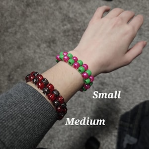 Könnte beinhalten: Zwei Perlenarmbänder am Handgelenk einer Person. Das obere Armband ist grün und pink und mit "Klein" beschriftet. Das untere Armband ist schwarz und rot und mit "Mittel" beschriftet.