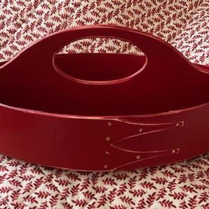 May include: A red oval-shaped wooden basket with a handle. The basket has a smooth, glossy finish and decorative white accents. The handle is a large oval shape, and the basket sits on a patterned fabric.