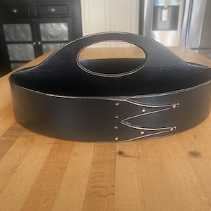 May include: A black oval-shaped wooden carrier with a central arched handle. The exterior features a distinct overlapping joint design secured with small copper-colored metal rivets, showing a subtle wood grain texture throughout the surface.