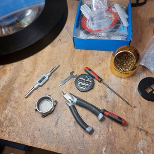 Peut inclure: Un ensemble d'outils et de composants de r&eacute;paration de montres, comprenant des pinces, des tournevis, un fer &agrave; souder et une loupe. Une bo&icirc;te contient des pi&egrave;ces de montre et du fil rouge. Les outils sont dispos&eacute;s sur une surface en bois.