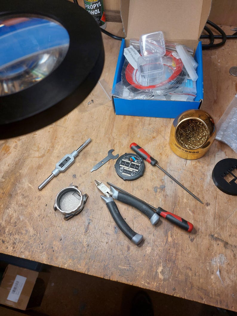 Peut inclure: Un ensemble d'outils et de composants de r&eacute;paration de montres, comprenant des pinces, des tournevis, un fer &agrave; souder et une loupe. Une bo&icirc;te contient des pi&egrave;ces de montre et du fil rouge. Les outils sont dispos&eacute;s sur une surface en bois.