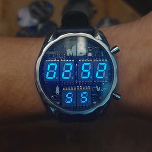 Op de afbeelding: Een futuristisch digitaal polshorloge met een zwarte leren band en een zilverkleurige kast. De wijzerplaat toont blauwe digitale cijfers. Het horloge heeft de letters "MB" op de wijzerplaat.