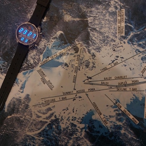 Peut inclure: Une montre num&eacute;rique avec un bracelet noir est pos&eacute;e sur une carte de la station de ski du Mont Buller. L'&eacute;cran de la montre affiche l'heure en chiffres bleus. La carte montre les pistes et les remont&eacute;es m&eacute;caniques.