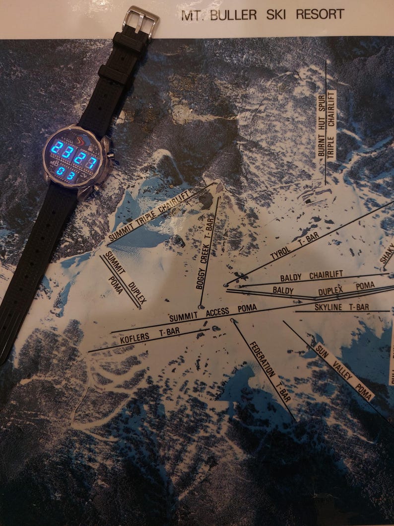 Peut inclure: Une montre num&eacute;rique avec un bracelet noir est pos&eacute;e sur une carte de la station de ski du Mont Buller. L'&eacute;cran de la montre affiche l'heure en chiffres bleus. La carte montre les pistes et les remont&eacute;es m&eacute;caniques.
