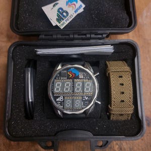 Peut inclure: Une montre num&eacute;rique avec un bracelet noir et un bo&icirc;tier argent&eacute; est pr&eacute;sent&eacute;e dans un &eacute;tui noir. Le cadran de la montre affiche des chiffres num&eacute;riques. Un deuxi&egrave;me bracelet de montre kaki et un autocollant avec le texte "TIME PIECES - BULLER" sont &eacute;galement visibles.