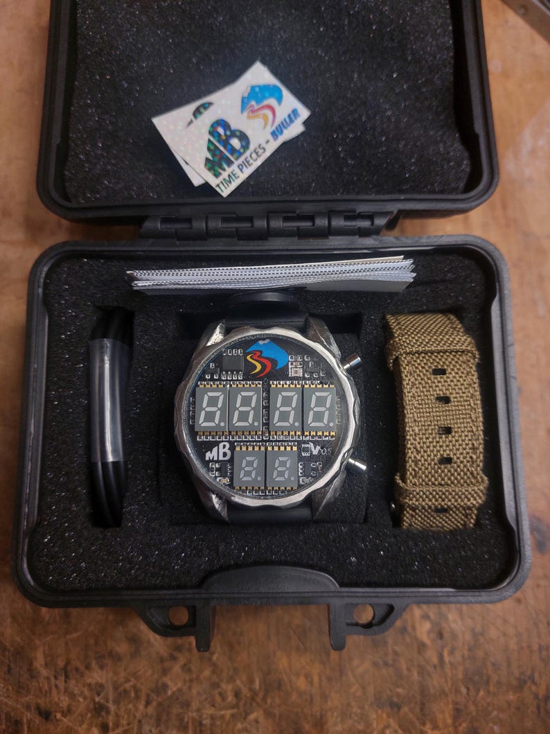 Peut inclure: Une montre num&eacute;rique avec un bracelet noir et un bo&icirc;tier argent&eacute; est pr&eacute;sent&eacute;e dans un &eacute;tui noir. Le cadran de la montre affiche des chiffres num&eacute;riques. Un deuxi&egrave;me bracelet de montre kaki et un autocollant avec le texte "TIME PIECES - BULLER" sont &eacute;galement visibles.