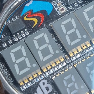 Peut inclure: Gros plan sur un cadran de montre avec un design de circuit imprim&eacute;. Le cadran pr&eacute;sente des affichages num&eacute;riques gris affichant des chiffres, un logo color&eacute; et divers composants &eacute;lectroniques. La montre a un bo&icirc;tier en m&eacute;tal argent&eacute;.