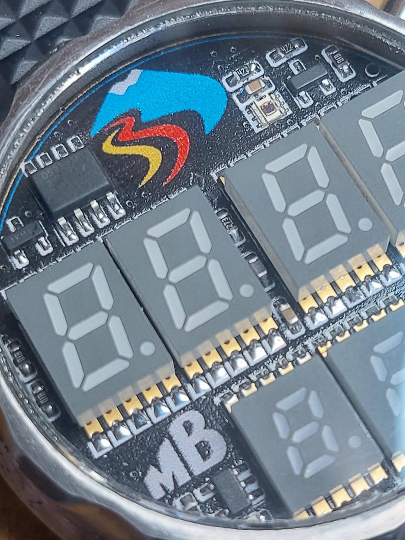Peut inclure: Gros plan sur un cadran de montre avec un design de circuit imprim&eacute;. Le cadran pr&eacute;sente des affichages num&eacute;riques gris affichant des chiffres, un logo color&eacute; et divers composants &eacute;lectroniques. La montre a un bo&icirc;tier en m&eacute;tal argent&eacute;.