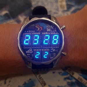 Peut inclure: Une montre-bracelet num&eacute;rique de style futuriste avec un bo&icirc;tier argent&eacute; et un bracelet noir. L'&eacute;cran affiche des chiffres et des lettres bleus vifs. La montre a un design unique avec plusieurs boutons et un logo.