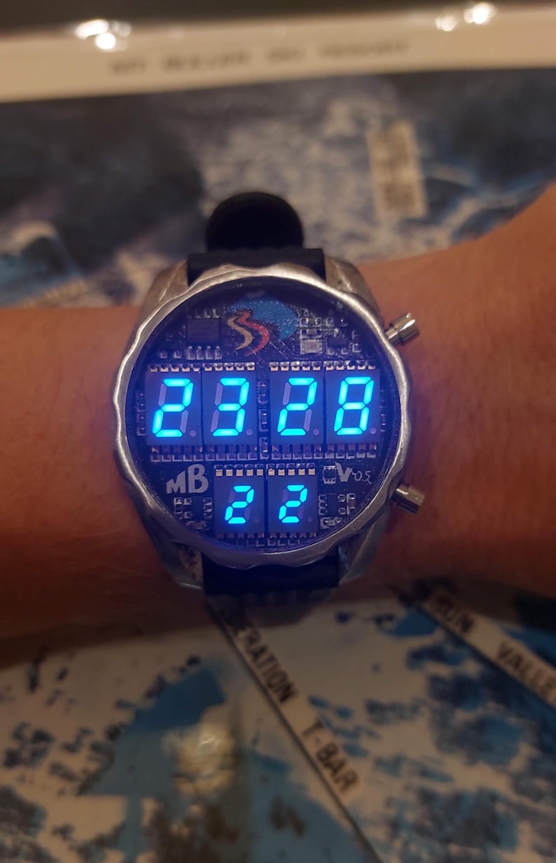 Peut inclure: Une montre-bracelet num&eacute;rique de style futuriste avec un bo&icirc;tier argent&eacute; et un bracelet noir. L'&eacute;cran affiche des chiffres et des lettres bleus vifs. La montre a un design unique avec plusieurs boutons et un logo.