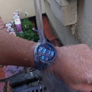Peut inclure: Une montre-bracelet num&eacute;rique avec un &eacute;cran bleu &eacute;clair&eacute; affichant l'heure. La montre est test&eacute;e sous l'eau courante. L'&eacute;cran affiche l'heure 23h35 et la date 5/1.