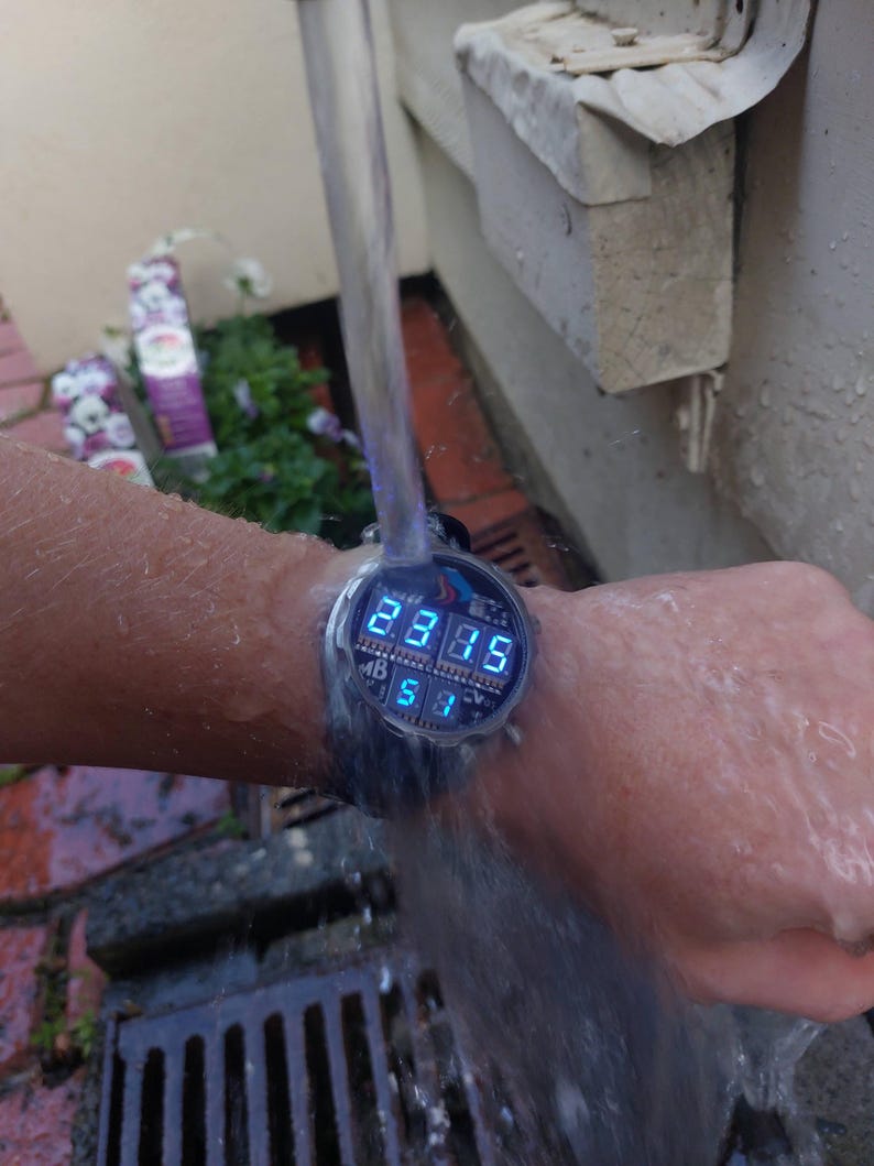 Peut inclure: Une montre-bracelet num&eacute;rique avec un &eacute;cran bleu &eacute;clair&eacute; affichant l'heure. La montre est test&eacute;e sous l'eau courante. L'&eacute;cran affiche l'heure 23h35 et la date 5/1.