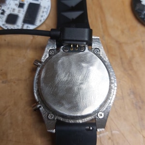 Peut inclure: Gros plan d'une montre argent&eacute;e avec un bracelet noir et un c&acirc;ble de chargement attach&eacute;. Le cadran de la montre est circulaire avec une finition textur&eacute;e. Le bracelet noir a un design textur&eacute;. Un circuit imprim&eacute; est visible en arri&egrave;re-plan.