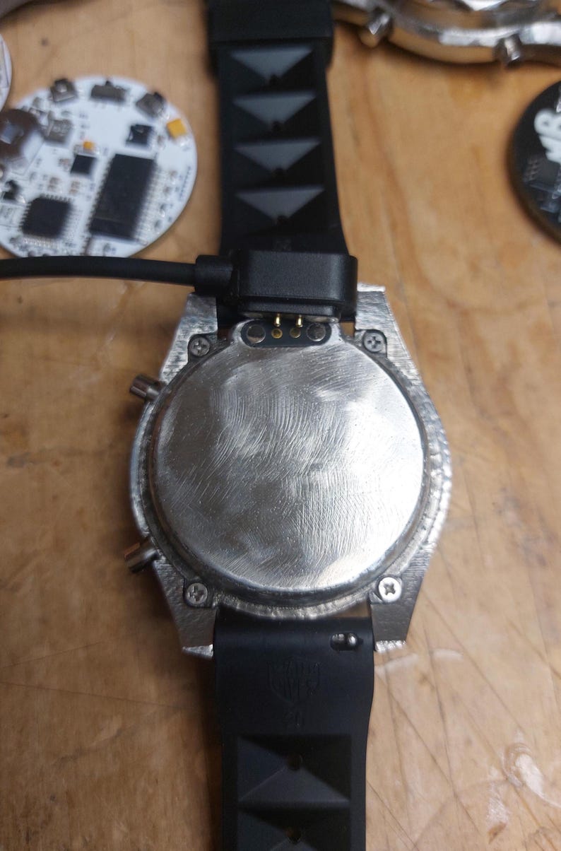 Peut inclure: Gros plan d'une montre argent&eacute;e avec un bracelet noir et un c&acirc;ble de chargement attach&eacute;. Le cadran de la montre est circulaire avec une finition textur&eacute;e. Le bracelet noir a un design textur&eacute;. Un circuit imprim&eacute; est visible en arri&egrave;re-plan.