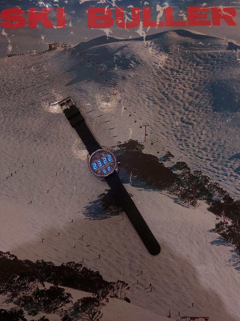 Peut inclure: Une montre-bracelet noire avec un cadran rond affichant l'heure num&eacute;rique, sur fond de paysage de montagne enneig&eacute;e. La montre a un bracelet noir et une boucle argent&eacute;e. Le fond pr&eacute;sente le texte "SKI BULLER" en rouge.