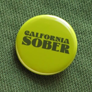 May include: A round, lime green button with the words "CALIFORNIA SOBER" printed in dark green. The button is set against a textured, dark green fabric background.