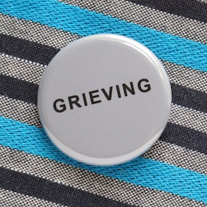 May include: A circular, pale grey badge with the word "GRIEVING" in black lettering. The badge is placed on a striped fabric with black, grey, and turquoise blue stripes.