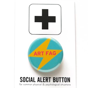 May include: A turquoise button with a yellow lightning bolt and the text "ART FAG" in pink. The button is on a white card with a black plus sign in a white square and the text "SOCIAL ALERT BUTTON for common physical & psychological situations".