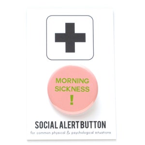 A round light pink pinback button that reads MORNING SICKNESS ! in puke green text. Pin is on a Social Alert Button backing card.