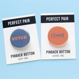 May include: Two pinback buttons on white cards with the text "PERFECT PAIR". One button is blue with "BUTCH" in orange, the other is orange with "FEMME" in blue. The cards read "PINBACK BUTTON" and "KEEP ONE" or "SHARE ONE".