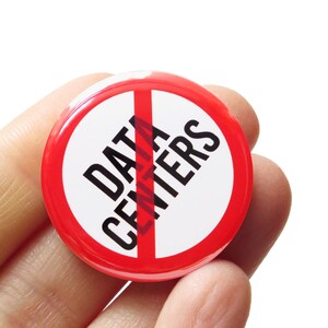May include: A round button with a red border and a white center. The text "DATA CENTERS" is printed in black, with a red line through it. The button is held in a hand.