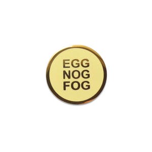 A small round yellowy cream colored enamel pin with a gold outline and three lines of gold text that reads, EGG NOG FOG.