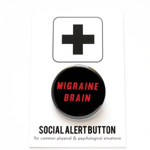 A round black pinback button that reads MIGRAINE BRAIN in red text. Badge is pinned to a Social Alert Button backing card, with a plus sign at the top.